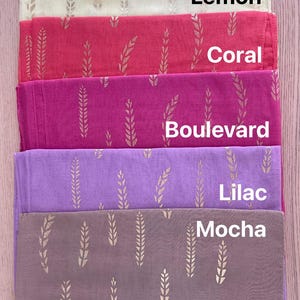 May include: A stack of five folded scarves in various colors: Lemon, Coral, Boulevard, Lilac, and Mocha. Each scarf features a gold wheat stalk pattern. The names of the colors are printed on the scarves.