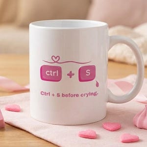 May include: White ceramic mug with a handle, featuring a pink design of "ctrl + S" keyboard keys and the phrase "Ctrl + S before crying." The mug is surrounded by pink candies and ribbons.