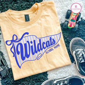 May include: A light yellow t-shirt featuring a blue pennant graphic with the text "Wildcats" and "SCHOOL NAME" in a script font. The pennant has a light blue vertical striped pattern and a blue ribbon accent on the left side of the design.