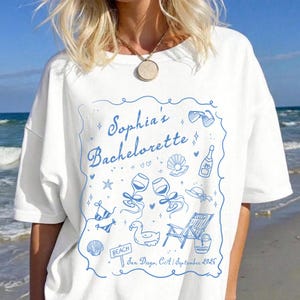 Może przedstawiać: Biała koszulka z niebieską ilustracją na wieczór panieński. Projekt zawiera napis "Sophia's Bachelorette" wraz z grafikami o tematyce plażowej, takimi jak okulary przeciwsłoneczne, kieliszki do wina i leżak. Zawiera również tekst "San Diego, CA | Wrzesień 2026".