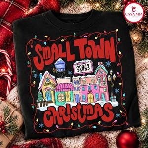 May include: A black sweatshirt featuring a graphic of a snowy town with colourful buildings, a water tower, and a sign reading 38863. The design is framed by red text reading Small Town Christmas and a string of multi-coloured Christmas lights.