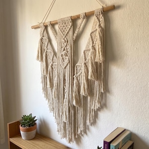 May include: Cream-colored macrame wall hanging with a geometric design. The wall decor features a wooden dowel at the top, with intricate knotting and tassels. The piece is suspended against a white wall, adding a touch of bohemian style to any space.