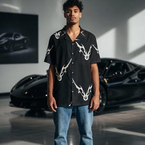 Pode incluir: Uma pessoa vestindo uma camisa preta com um design tribal branco, jeans azuis e sapatos pretos. Um carro esportivo preto é visível ao fundo. A cena se passa em um showroom.