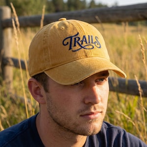 May include: Mustard yellow baseball cap with the word "TRAILS" embroidered in navy blue script across the front. The cap has a curved brim and a classic six-panel design. The hat is made of a soft, worn material.