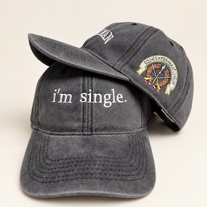 May include: Two charcoal grey baseball caps stacked together. The front cap features the white embroidered text 'i'm single.' The top cap displays a circular embroidered patch with a spear, crossed rifles, and the Latin text 'ECO SEMPER PARATUS SUM'.
