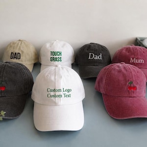 May include: A collection of baseball caps in various colours including beige, white, charcoal, and maroon. The caps feature embroidered text such as DAD, TOUCH GRASS, Dad, Mum, and Custom Logo Custom Text, along with small red cherry embroidery designs.
