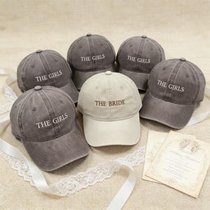 May include: A collection of six baseball caps. Five are gray with "THE GIRLS" embroidered in white, and one is beige with "THE BRIDE" in brown. The caps are arranged with decorative ribbons and a wedding invitation.
