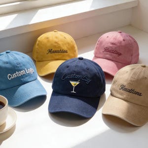 May include: Five baseball caps in shades of blue, yellow, pink, navy, and tan. Each cap has embroidered text or a logo. A cup of coffee is visible in the foreground.