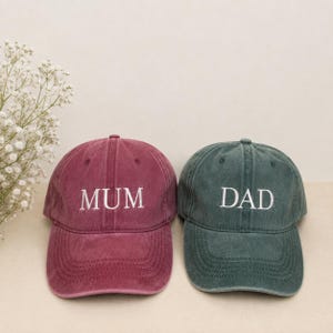 May include: A maroon baseball cap with the word MUM embroidered in white and a dark teal baseball cap with the word DAD embroidered in white. Both caps feature a structured crown, a curved brim, and a washed fabric finish with visible stitching.