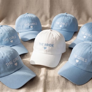 May include: A collection of light blue and white baseball caps. The light blue caps have "THE GIRLS" embroidered in white with a heart. The white cap says "THE BRIDE" with a blue heart. Perfect for a bridal party.