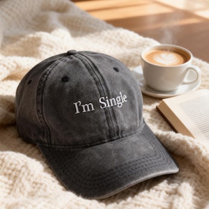 May include: A charcoal gray baseball cap with the embroidered text "I'm Single" in white. The hat is sitting on a cream-colored knitted blanket, with a cup of coffee and a book in the background.