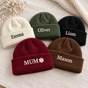 May include: Five knit beanies in cream, olive green, black, burgundy, and brown. Each hat features a folded cuff with embroidered text: Emma, Oliver, Liam, Mason, and MUM with a small white daisy flower icon next to the word MUM.