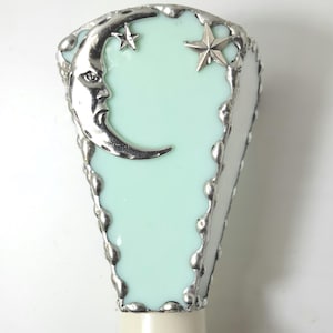 May include: A triangular-shaped light fixture shade featuring a pale mint green glass panel framed in textured silver metal solder. The front surface is decorated with a silver crescent moon with a face and two silver stars of different sizes.