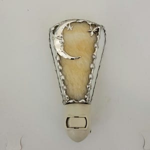 May include: A plug-in night light featuring a tapered, cream-colored textured glass shade framed in silver metal. The front panel displays a silver crescent moon with a face and two small silver stars. A white plastic base includes an on-off toggle switch.