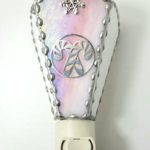 May include: A stained glass night light featuring a pale pink and iridescent glass panel with a silver snowflake accent at the top and a circular silver candy cane emblem in the center. The glass is framed in silver solder with a beaded edge.