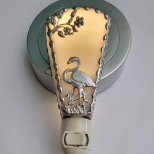 May include: A decorative night light featuring a silver-colored flamingo design. The night light has a warm, soft glow and is framed by a silver-colored border with floral accents. The base is off-white and the light is plugged into a silver-colored wall outlet.