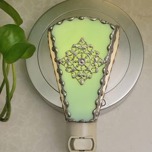 May include: A green glass night light with a silver filigree design. The night light has a white plug-in base with a switch.