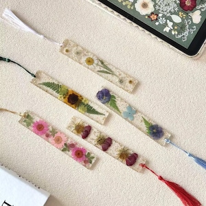 Pode incluir: Uma coleção de marcadores de livros retangulares de resina, cada um com flores secas embutidas. Os marcadores de livros apresentam vários arranjos florais, incluindo margaridas, girassóis e outras flores coloridas. Cada marcador tem uma borla.