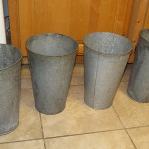 May include: Four galvanized metal buckets with a tapered design. The buckets are a dull gray color and appear to be vintage or rustic. They are arranged in a row on a tiled floor, with a wooden cabinet in the background.