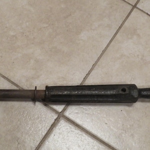 May include: A dark-coloured vintage tool with a curved claw at one end and a handle at the other. The tool appears to be made of metal and has a cylindrical body. The handle is black and rounded.