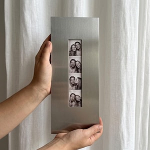 Puede incluir: Un marco de fotos rectangular de metal cepillado con un recorte vertical en el centro. El marco muestra una tira vertical de cuatro fotografías en blanco y negro que muestran a una pareja haciendo varias expresiones faciales al estilo fotomatón.