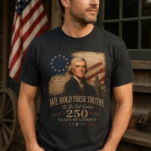 May include: Dark gray t-shirt featuring a patriotic design with the text "We Hold These Truths" and an image of Thomas Jefferson. The design includes an American flag and the text "250 Years of Liberty."