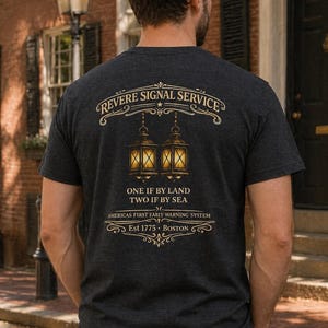 May include: Dark gray t-shirt with gold lettering and graphics. The shirt features the text "REVERE SIGNAL SERVICE" above a design of two lanterns. Below the lanterns, the text reads "ONE IF BY LAND TWO IF BY SEA" and "AMERICA'S FIRST EARLY WARNING SYSTEM".