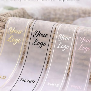 Könnte beinhalten: Weiße Bänder mit dem Text "Your Logo" in Gold, Silber, Schwarz, Weiß und Rosa bedruckt. Die Bänder werden mit einem Weidenkorb im Hintergrund präsentiert. Der Text "Your Logo with Many Print Color Options" steht oben.
