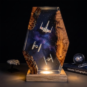 May include: A hexagonal lamp featuring a central resin panel with a deep space nebula background, an X-wing starfighter, and two TIE fighters. The panel is framed by two pieces of natural live-edge wood and sits on a rectangular wooden base.