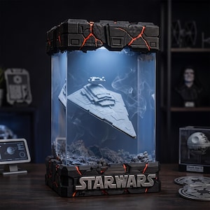 May include: A Star Wars display case featuring a grey Imperial Star Destroyer model suspended inside a clear rectangular enclosure. The base and top are black with glowing orange cracks, and the base displays the silver text STAR WARS.