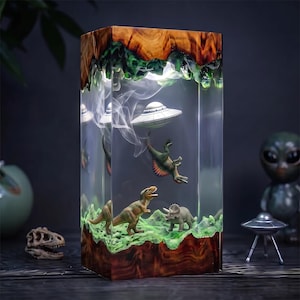 Può includere: Una lampada rettangolare in resina con base e parte superiore in legno naturale, che racchiude una scena in miniatura con un UFO argentato fluttuante, uno Spinosaurus sospeso, un Tyrannosaurus Rex e un Triceratops su un paesaggio verde.