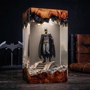 May include: A rectangular lamp featuring a Batman figurine standing on a metallic bridge structure inside a clear resin block. The top and bottom sections are made of natural wood with a dark grain, and the interior is illuminated by warm light.