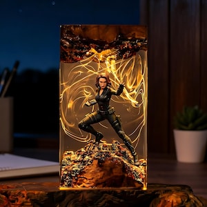 Puede incluir: Una lámpara rectangular de resina con una figura de Black Widow en pose de combate dinámica. La figura se alza sobre una base rocosa texturizada dentro de un bloque transparente, rodeada de patrones de luz dorada y detalles de madera.