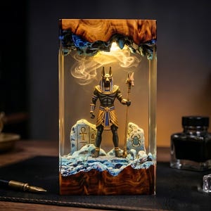 May include: A rectangular resin lamp featuring a central Anubis figurine holding a staff. The base and top are made of natural wood with blue accents. The interior is illuminated, casting a warm glow over the figure and two stone-like carvings.