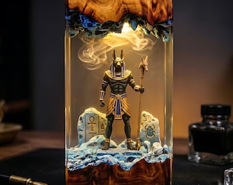 May include: A rectangular resin lamp featuring a central Anubis figurine holding a staff. The base and top are made of natural wood with blue accents. The interior is illuminated, casting a warm glow over the figure and two stone-like carvings.