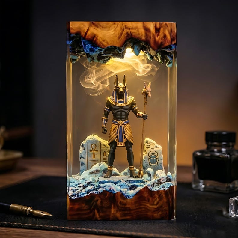 May include: A rectangular resin lamp featuring a central Anubis figurine holding a staff. The base and top are made of natural wood with blue accents. The interior is illuminated, casting a warm glow over the figure and two stone-like carvings.