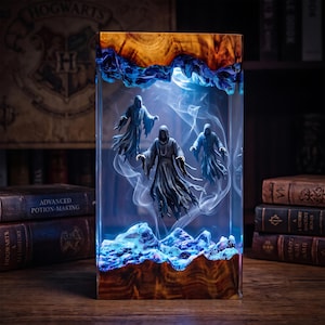 May include: A rectangular lamp featuring a natural wood base and top with a clear resin center. Inside the resin, three hooded figures in flowing robes appear to float amidst swirling white smoke, illuminated by a cool blue internal light.