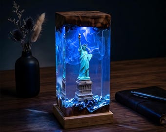 May include: A decorative lamp featuring the Statue of Liberty encased in a clear resin block. The statue is a teal color, set against a blue, swirling background. The lamp has a wooden base and top, creating a unique home decor item.