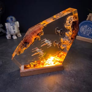 May include: A rectangular resin lamp featuring embedded wooden elements and miniature starfighter models. The clear resin displays two X-wing fighters and two TIE fighters in a space scene, illuminated from below by a warm orange light base.