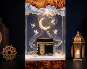 May include: A rectangular resin lamp featuring a wooden base and top, enclosing a miniature black Kaaba structure. Inside, a glowing crescent moon hangs above the Kaaba, surrounded by three white doves and swirling white smoke-like resin effects.