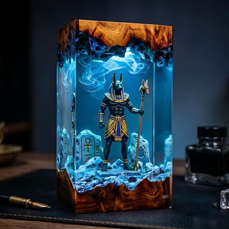 May include: A rectangular resin lamp featuring a wooden base and top, enclosing a miniature Anubis figure holding a staff. The interior is illuminated with a bright blue light, creating a smoky effect around the figure and stone-like carvings.