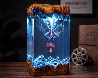 May include: A rectangular lamp featuring a natural wood base and top with a clear resin center. Inside the resin, a miniature red and blue superhero figure hangs upside down, suspended by a glowing blue light effect that resembles a web.