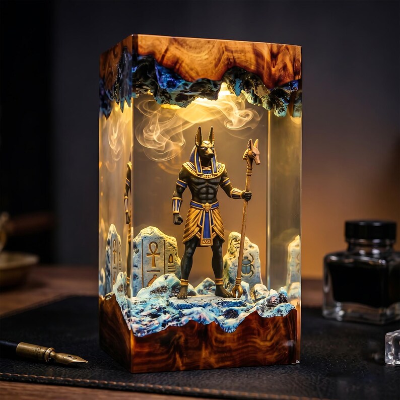 May include: A rectangular resin and wood lamp featuring an illuminated interior with a miniature Anubis figure holding a staff. The scene includes stone-like carvings with ankh and scarab symbols, set against a warm glowing light effect.