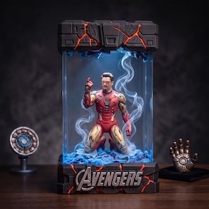 Puede incluir: Una figura coleccionable de Iron Man con traje rojo y dorado, arrodillada dentro de una vitrina rectangular con efectos de luz azul y humo. La base presenta la palabra AVENGERS en letras plateadas con grietas de color naranja brillante.