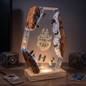 May include: A hexagonal lamp featuring a central Millennium Falcon and two TIE fighters suspended in clear resin between two jagged pieces of natural wood with white textured edges. The lamp sits on a rectangular wooden base with internal lighting.