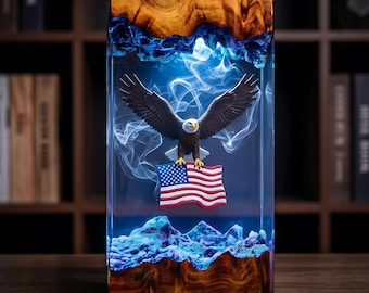 May include: A rectangular resin and wood sculpture featuring a bald eagle with wings spread wide, clutching an American flag. The piece is illuminated with a blue light, creating a swirling smoke effect between the natural wood top and base.