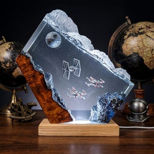 Peut inclure: Une lampe rectangulaire en résine représentant une scène spatiale avec une Étoile de la Mort, un chasseur TIE et deux chasseurs X-wing incrustés. L'objet présente un bord en bois naturel, une base en résine bleue texturée et un socle en bois clair.