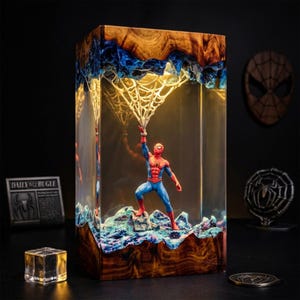 Op de afbeelding: Een rechthoekige lamp van hout en heldere hars met een Spider-Man-figuur op een getextureerde basis. Het figuur houdt een witte webstructuur vast die tot aan de bovenkant reikt. Het hout heeft een natuurlijke nerf en de hars bevat blauwe accenten.