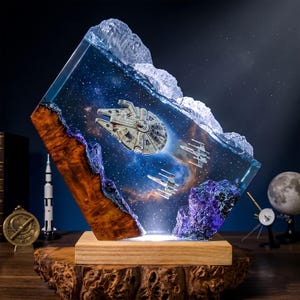 May include: A decorative resin and wood lamp featuring a space scene with a Millennium Falcon and two X-wing starfighters embedded within a clear, starry nebula background. The piece is mounted on a natural wood base with a textured, organic edge.