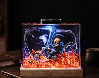 May include: A rectangular resin block lamp featuring a miniature figure of a flaming skull character riding a motorcycle. The scene is set amidst orange and red flames with swirling blue and white smoke effects, all mounted on a wooden base.